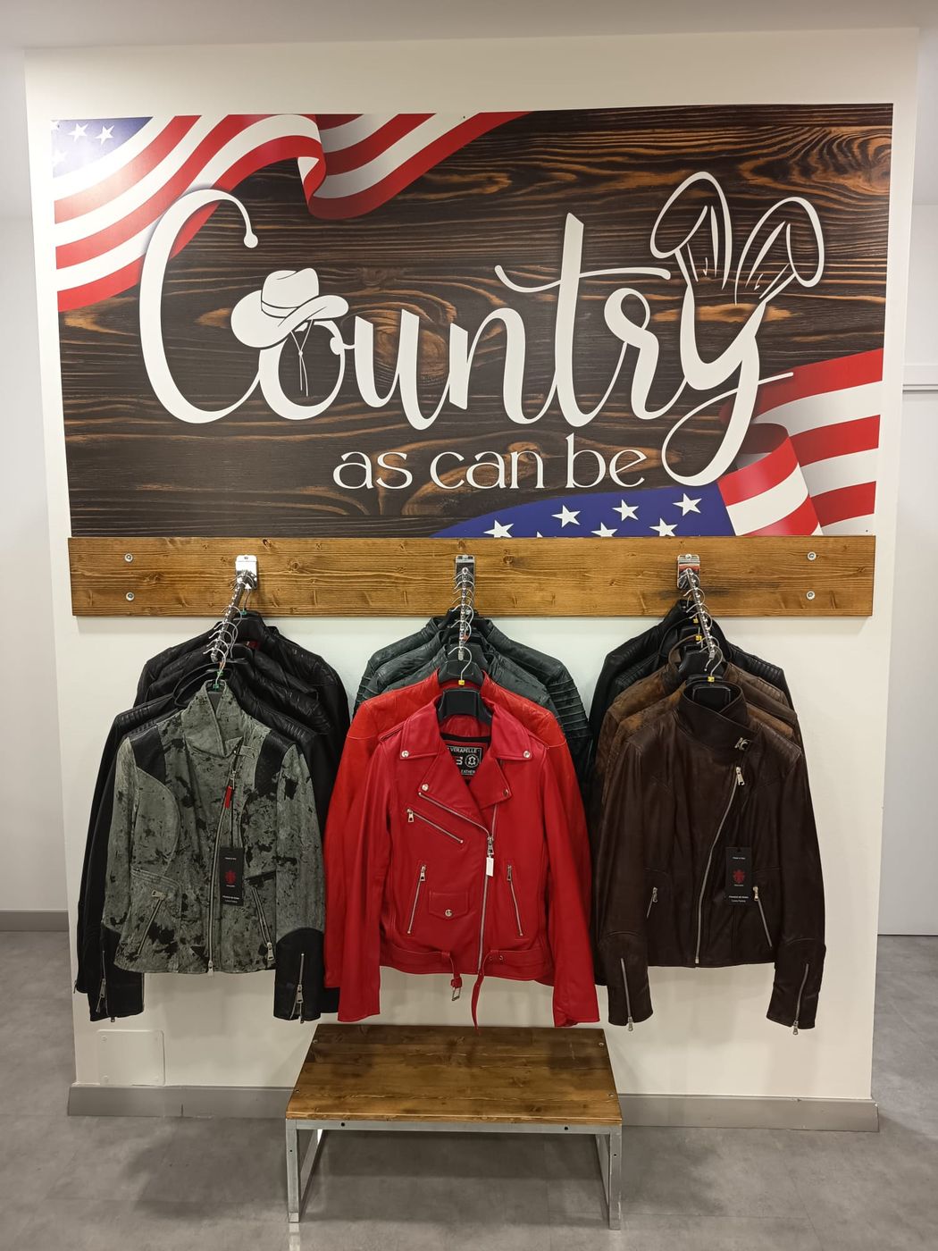 Parete interna del negozio con l’insegna Country as can be su fondo di legno e bandiera americana, e tre giacche in pelle appese sotto